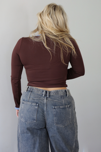 A woman shown from behind wearing a dark brown, fitted, long sleeved, slightly cropped top with blue trim detailing at the v-neckline and at the sleeve hems, paired with dark acid washed barrel denim jeans, standing indoors in front of a white wall.