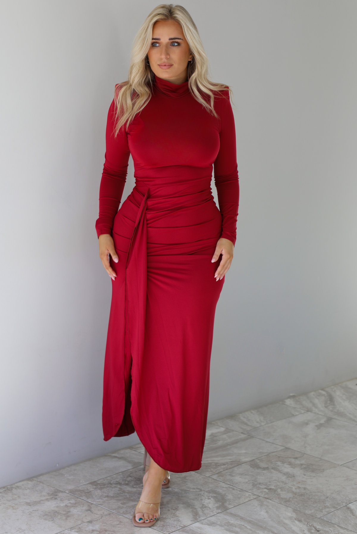 Woman wearing a red maxi dress that has a high neckline, shoulder pads, a fitted bodice, long sleeves, with a split hem detail on the left side. The photo is taken indoors against a white wall with white marble flooring. 