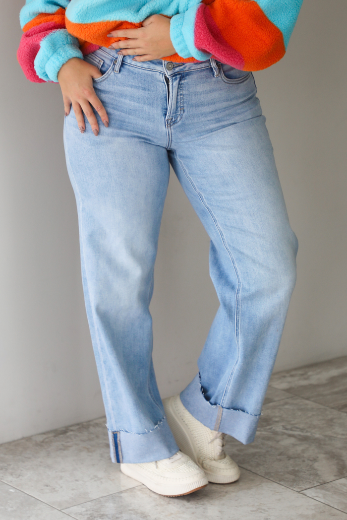 A woman shown from the waist down wearing a pair of light wash denim jeans with a cuffed hem, standing indoors against a white wall with white marble flooring. 