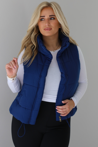 Woman wearing a navy puffer vest, white long-sleeve shirt, and black leggings indoors against a white wall. The puffer vest has a collared design, functional button closures at the neckline, an unzipped closure in the front, and functional pockets on either side.