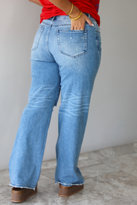 A woman shown from behind wearing light-wash blue distressed jeans with a faded wash, paired with a red t-shirt and brown platform sandals, standing indoors against a white wall with white marble flooring. 