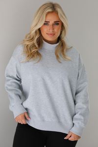 Woman wearing a grey turtleneck sweater, untucked, with high waisted black leggings. The photo is taken indoors against a white wall.