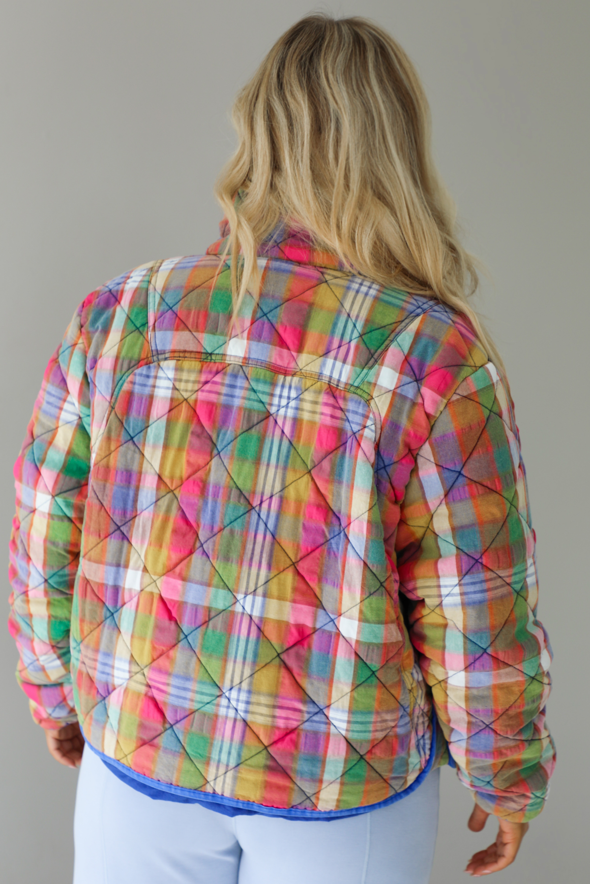 Woman wearing a multi-colored plaid, quilted style jacket paired with a white top underneath and high waisted blue lounge pants. The photo shows a closeup of the back of the jacket. The photo is taken indoors against a white wall.