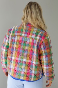 Woman wearing a multi-colored plaid, quilted style jacket paired with a white top underneath and high waisted blue lounge pants. The photo shows a closeup of the back of the jacket. The photo is taken indoors against a white wall.