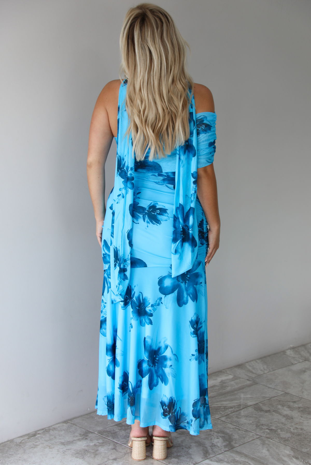 Woman wearing a blue floral maxi dress with a fitted bodice and flowy skirt indoors against a white wall with white marble flooring. The photo shows the back of the dress. 