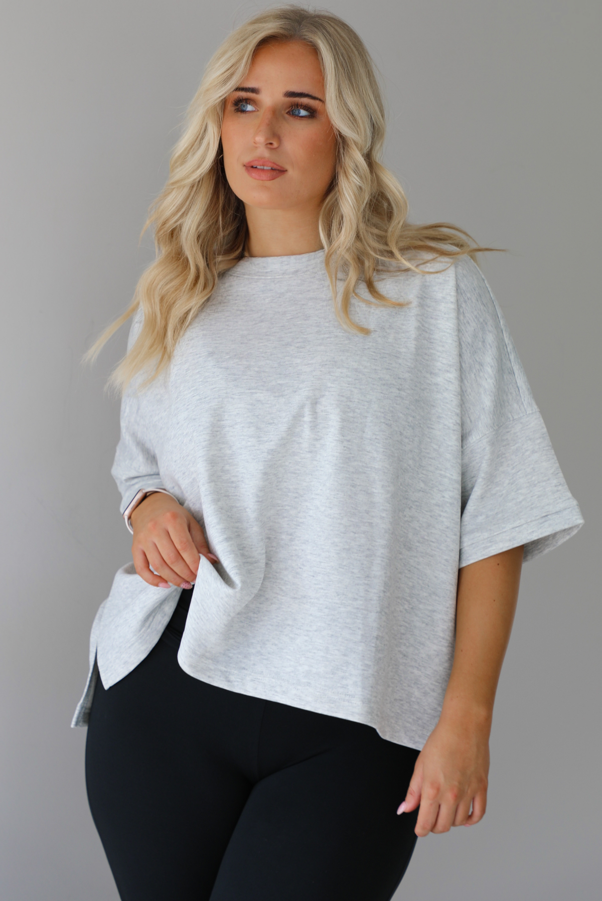 Woman wearing an oversized heather grey box tee with black leggings that flare slightly at the hems. The photo shows a closeup of the tee. The photo is taken indoors against a white wall.