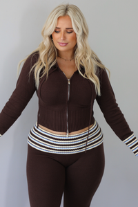 A woman modeling a dark brown ribbed zip-up hoodie and matching flared pants set with white striped accents on the cuffs and waistband, focused on the top, standing indoors against a white wall.
