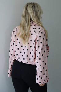 A woman shown from behind wearing a pink button-down jacket with a black polka dot print pattern, paired with black leggings, white ankle socks, and black slide on slippers, standing indoors against a white wall.