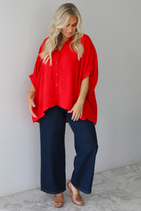 A woman wearing a bright red oversized short-sleeve blouse and dark wide-leg jeans, standing indoors against a white wall with white marble flooring. 