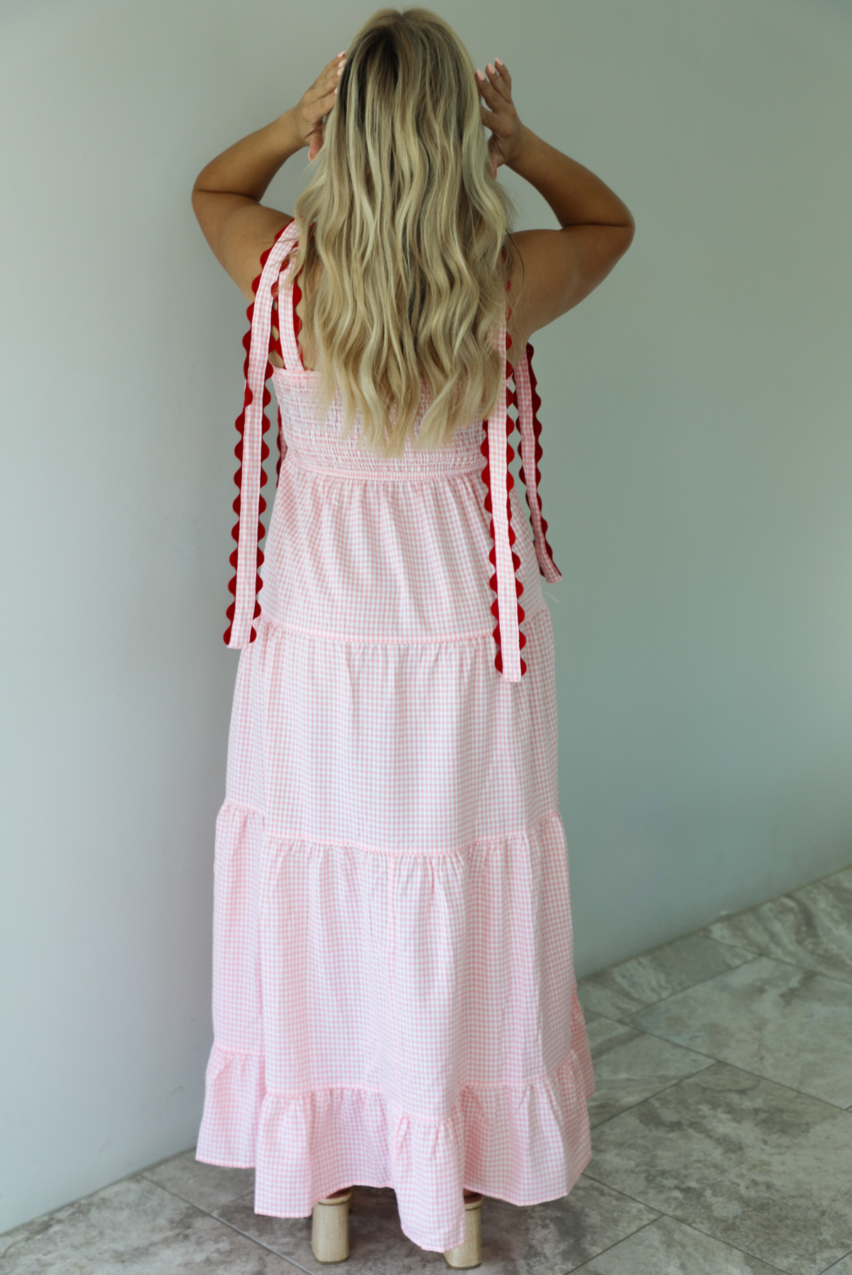 Woman wearing a pink and white gingham  maxi dress with thick straps, a fitted bodice, flowy skirt, and crocheted cherry details on the front indoors against a white wall with white marble flooring. The photo shows the back of the dress, which has a smocked detail at the waistline. 