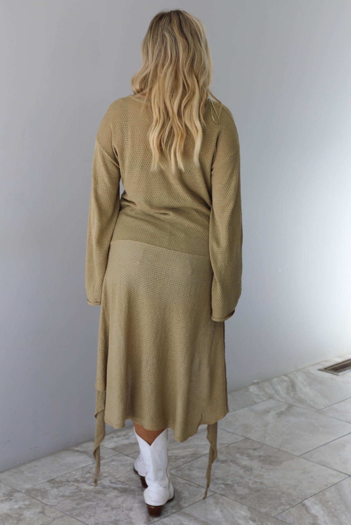 Woman wearing a camel long-sleeve dress that has a tie detail in the front.  The photo is taken indoors against a white wall. The photo shows the back of the dress. 