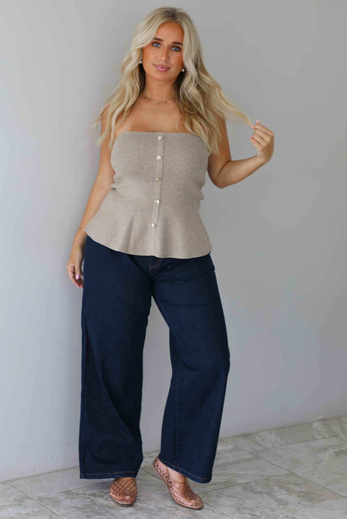 A woman wearing a beige strapless button-front peplum top and dark denim wide-leg jeans, standing indoors against a white wall with white marble flooring. 
