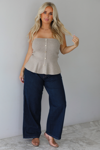 A woman wearing a beige strapless button-front peplum top and dark denim wide-leg jeans, standing indoors against a white wall with white marble flooring. 