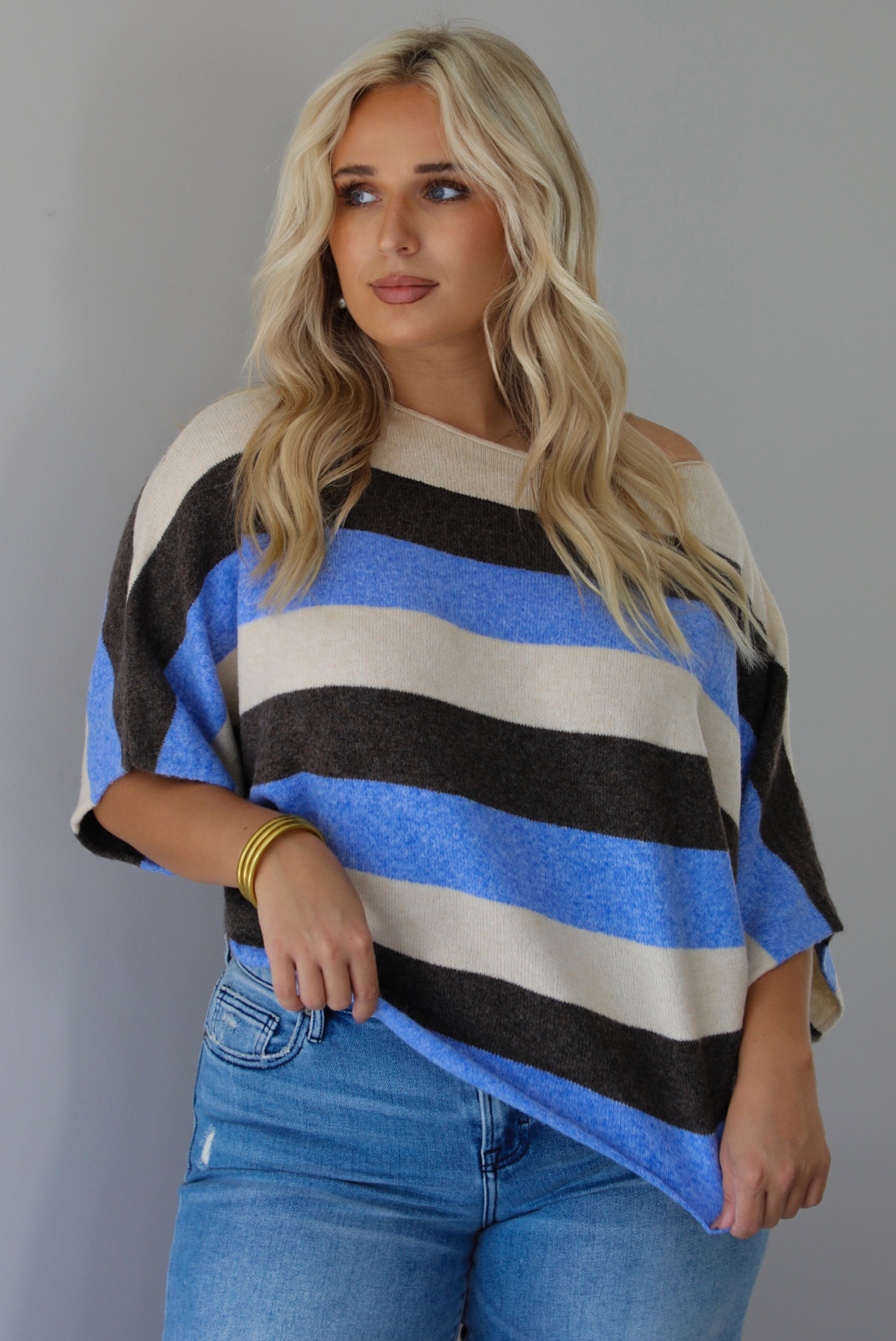 A woman wearing a cream, brown,, and blue striped knit top that has a boat-neck design, and quarter length flowy sleeves, paired with a pair of medium wash denim jeans, standing indoors against a white wall.