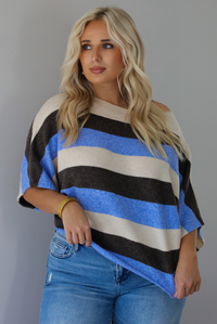 A woman wearing a cream, brown,, and blue striped knit top that has a boat-neck design, and quarter length flowy sleeves, paired with a pair of medium wash denim jeans, standing indoors against a white wall.