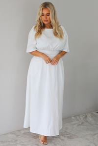 A woman modeling a white midi dress with short puff sleeves, a wide boat neckline, and side pockets, standing indoors against a white wall with white marble flooring. 