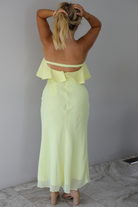 A woman viewed from behind wearing a pale yellow ruffled strapless maxi dress, standing indoors against a white wall with white marble flooring. 