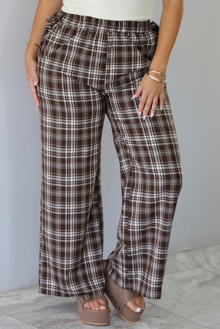 A person wearing brown plaid wide-leg pants with a white and dark stripe pattern, paired with a white top and brown platform sandals, shown from the waist down indoors against a white wall with white marble flooring. 