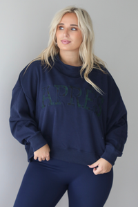 A woman wearing a navy sweater with Apreś written on the front in collegiate lettering in a plaid print, paired with a matching two piece navy set, standing indoors against a white wall.