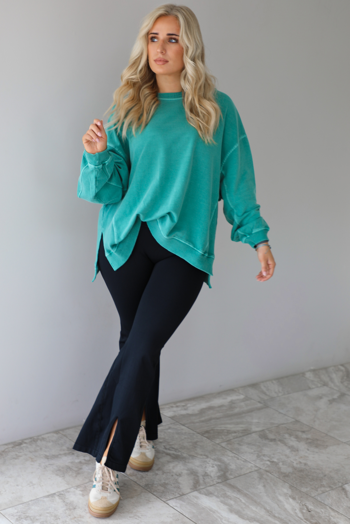 Woman wearing a teal oversized sweater with black leggings that have a slightly flared hem with split hem details in the front. The photo is taken in front of a white wall with white marble flooring. 