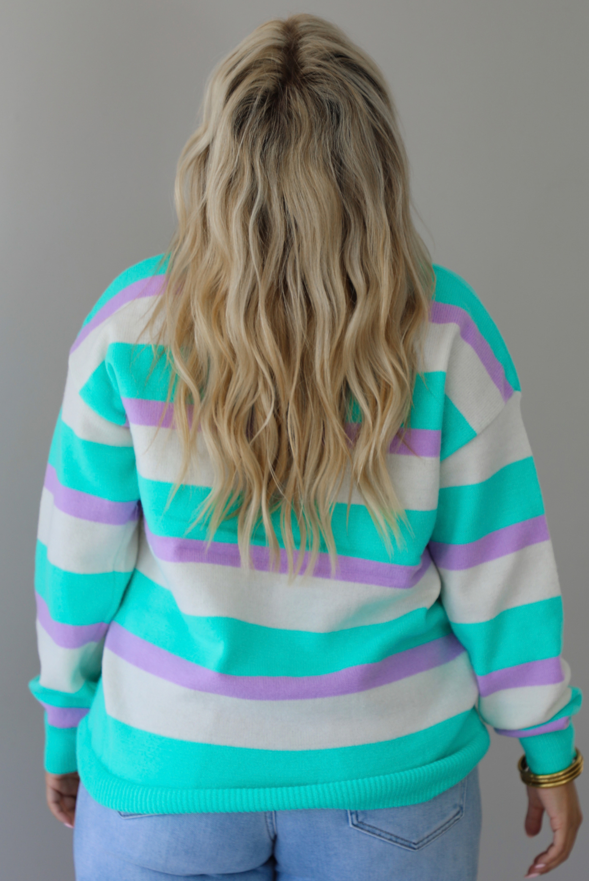 A woman shown from behind wearing a turquoise, lavender, and cream striped knit sweater, untucked to show the full length, paired with light wash denim jeans, standing indoors against a white wall.