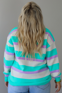 A woman shown from behind wearing a turquoise, lavender, and cream striped knit sweater, untucked to show the full length, paired with light wash denim jeans, standing indoors against a white wall.