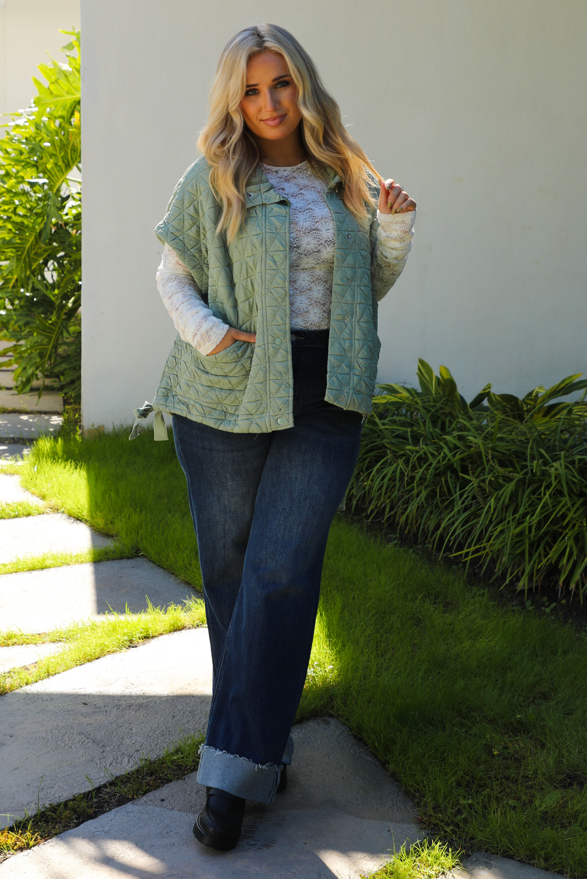 Woman model wearing a sage colored oversized vest paired with a white lace long sleeved top and dark denim outdoors.  The vest has functional pockets on either side, functional button closures in the front, and an oversized fit. 