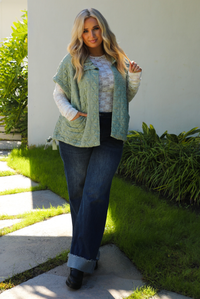 Woman model wearing a sage colored oversized vest paired with a white lace long sleeved top and dark denim outdoors.  The vest has functional pockets on either side, functional button closures in the front, and an oversized fit. 