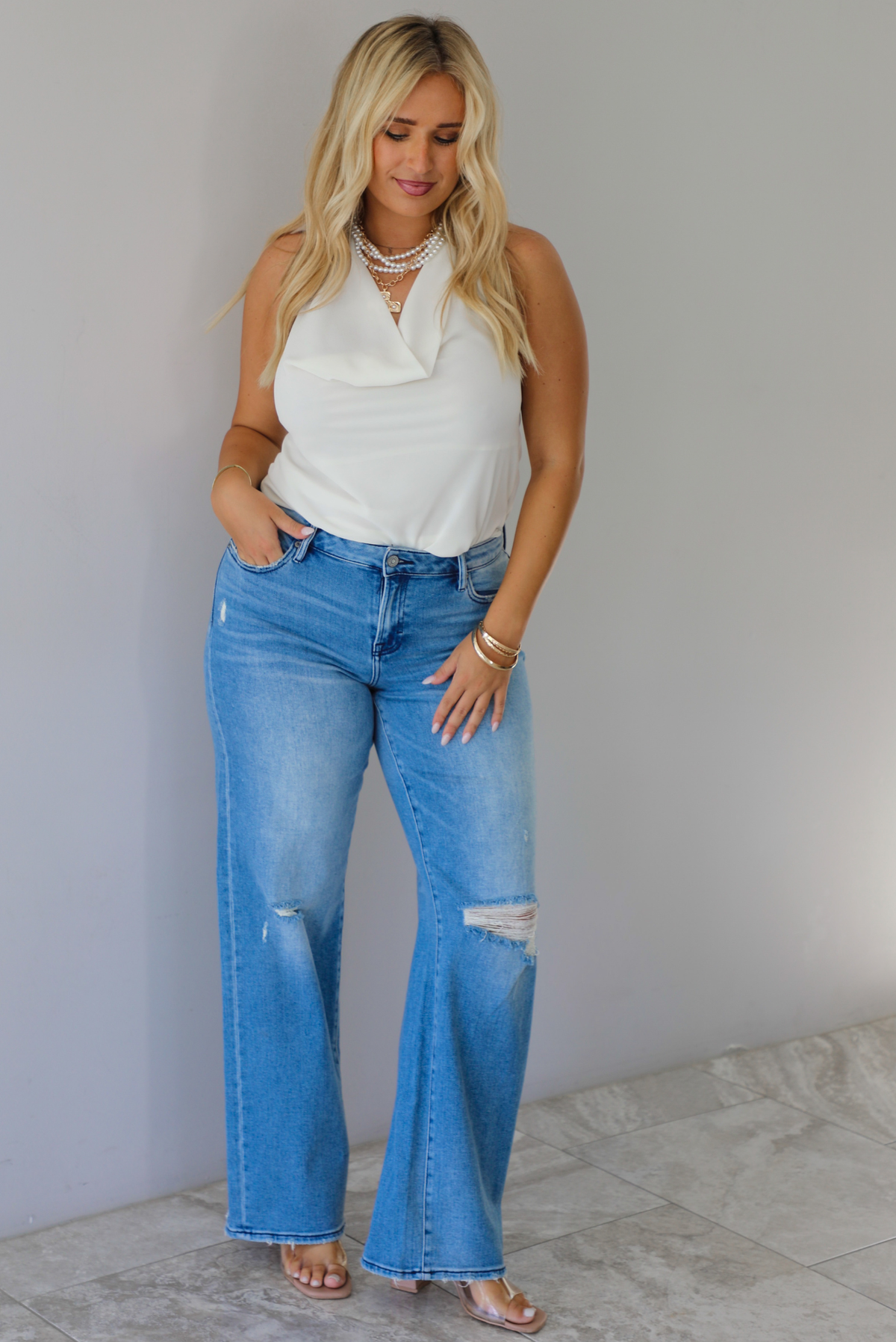 A woman modeling a cream sleeveless halter top and light-wash blue distressed flare jeans, standing indoors against a white wall with white marble flooring. 