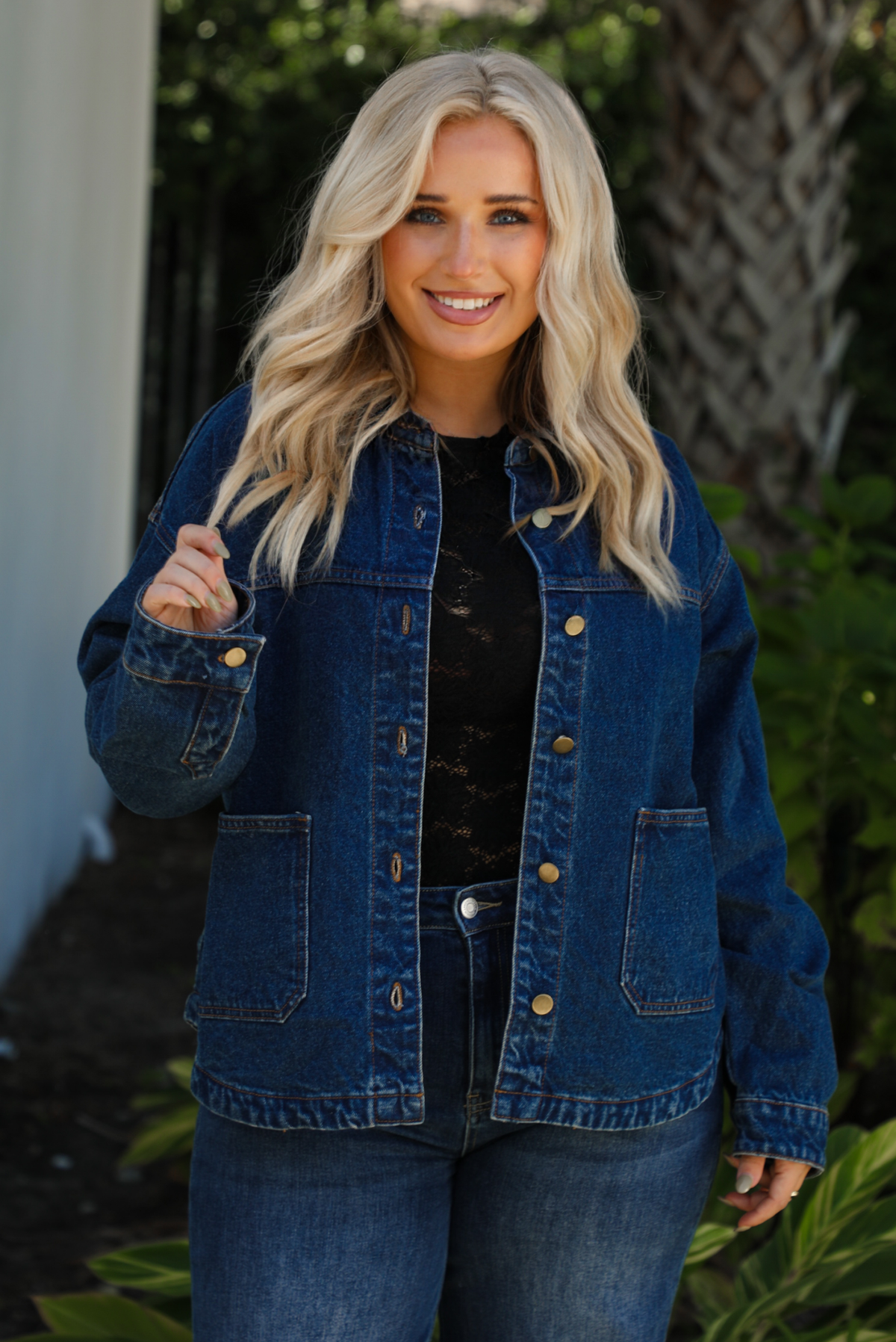 Close up of a woman model wearing a dark denim button up jacket showcasing the button closures in the front along with the pockets, paired with a black lace top,  and dark denim jeans outdoors. 