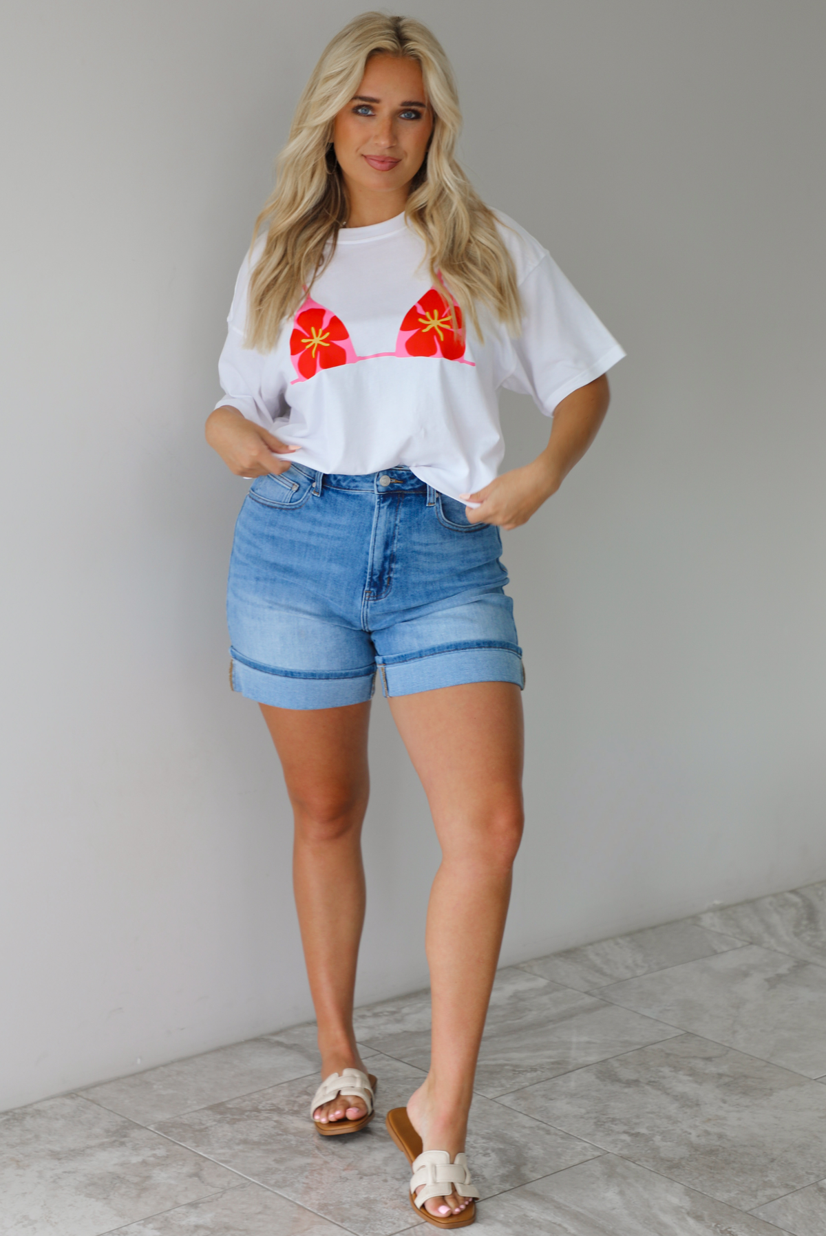 Woman wearing a white cropped boxy tee with a pink, red, and yellow bikini graphic on the front with a pair of medium wash denim shorts indoors against a white wall with white marble flooring.