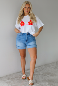 Woman wearing a white cropped boxy tee with a pink, red, and yellow bikini graphic on the front with a pair of medium wash denim shorts indoors against a white wall with white marble flooring.