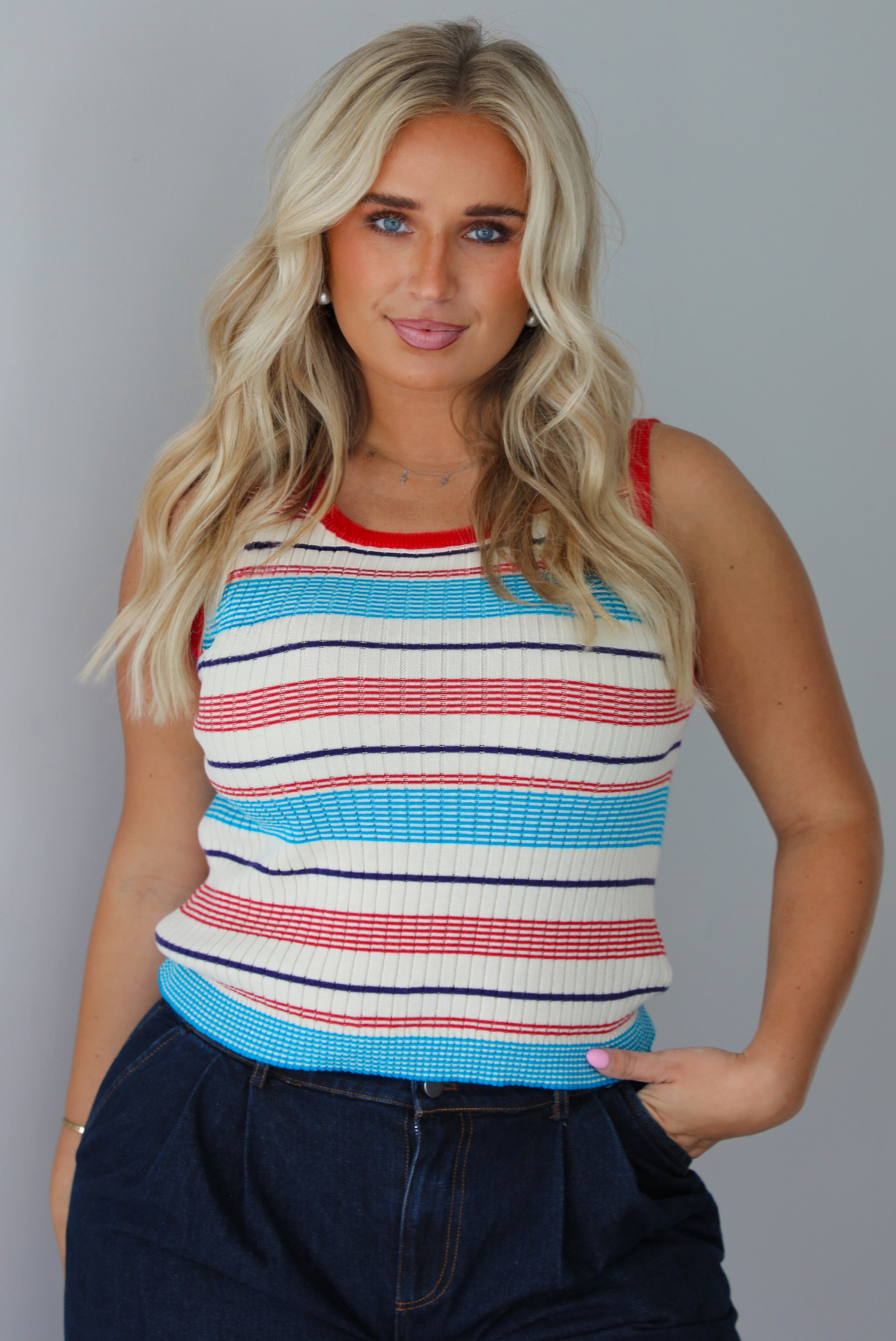 A woman modeling a sleeveless ribbed knit top with horizontal stripes in red, light blue, navy, and white, paired with dark wide-leg jeans and metallic rose gold flats, standing indoors against a white wall.