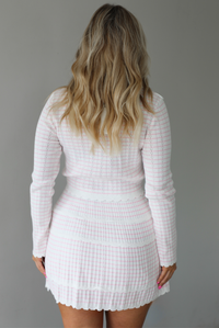 Woman wearing a two piece set that featuring a cropped cardigan and a high waisted skirt. The set is white with a pink horizontal striped pattern throughout, with scalloped hem details. The photo shows the back of the set. The photo is taken indoors against a white wall.