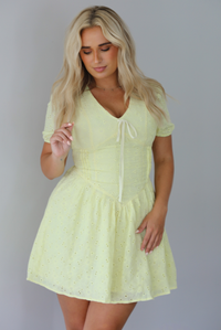 Woman wearing a light yellow mini dress with a floral eyelet design throughout, v-neckline, short sleeves, fitted drop waist bodice, and a flowy mini length skirt indoors against a white wall.