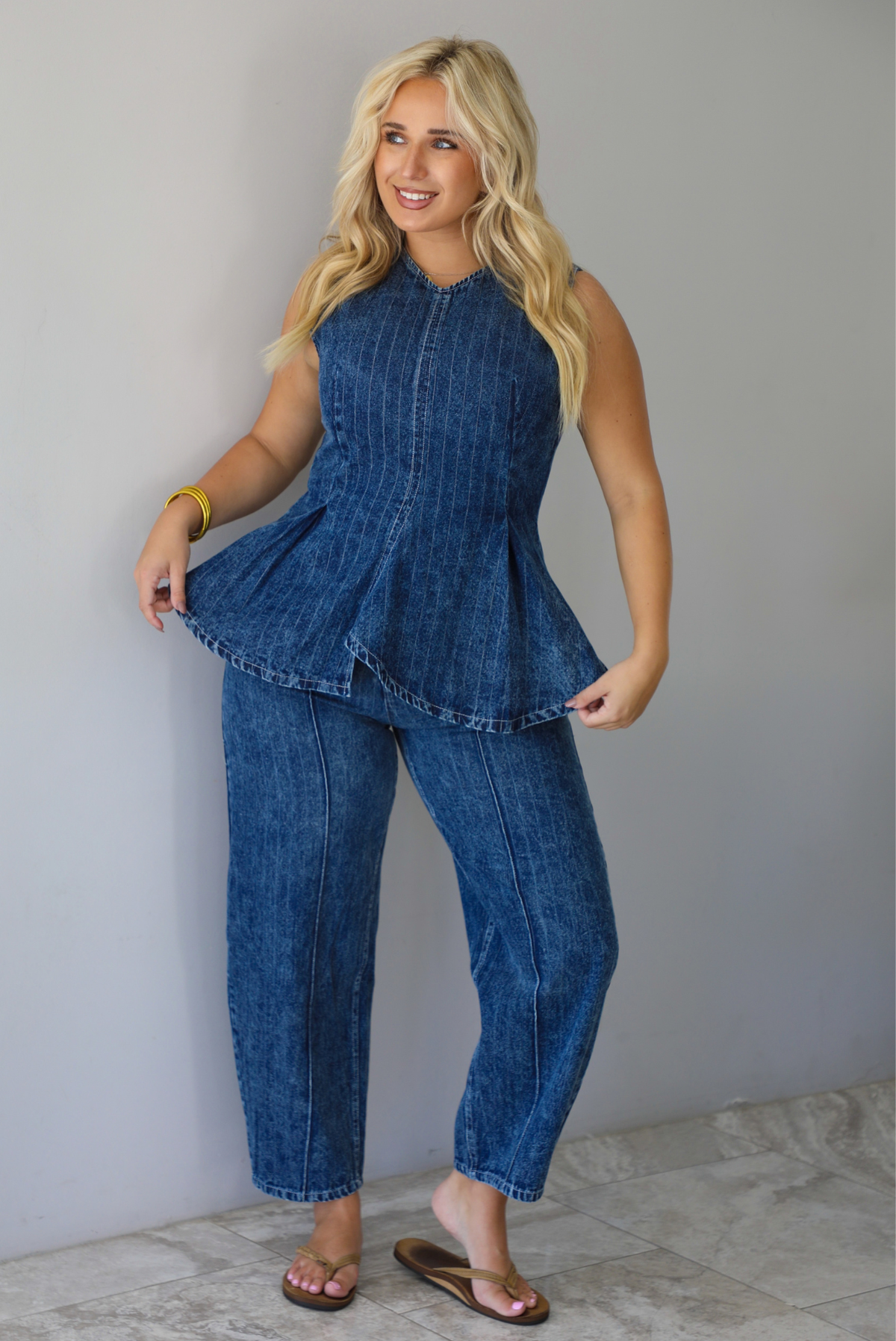 A woman wearing a matching two piece dark wash denim set with a pin striped design on it featuring a sleeveless peplum style top and high waisted denim jeans, standing indoors against a white wall with white marble flooring. 