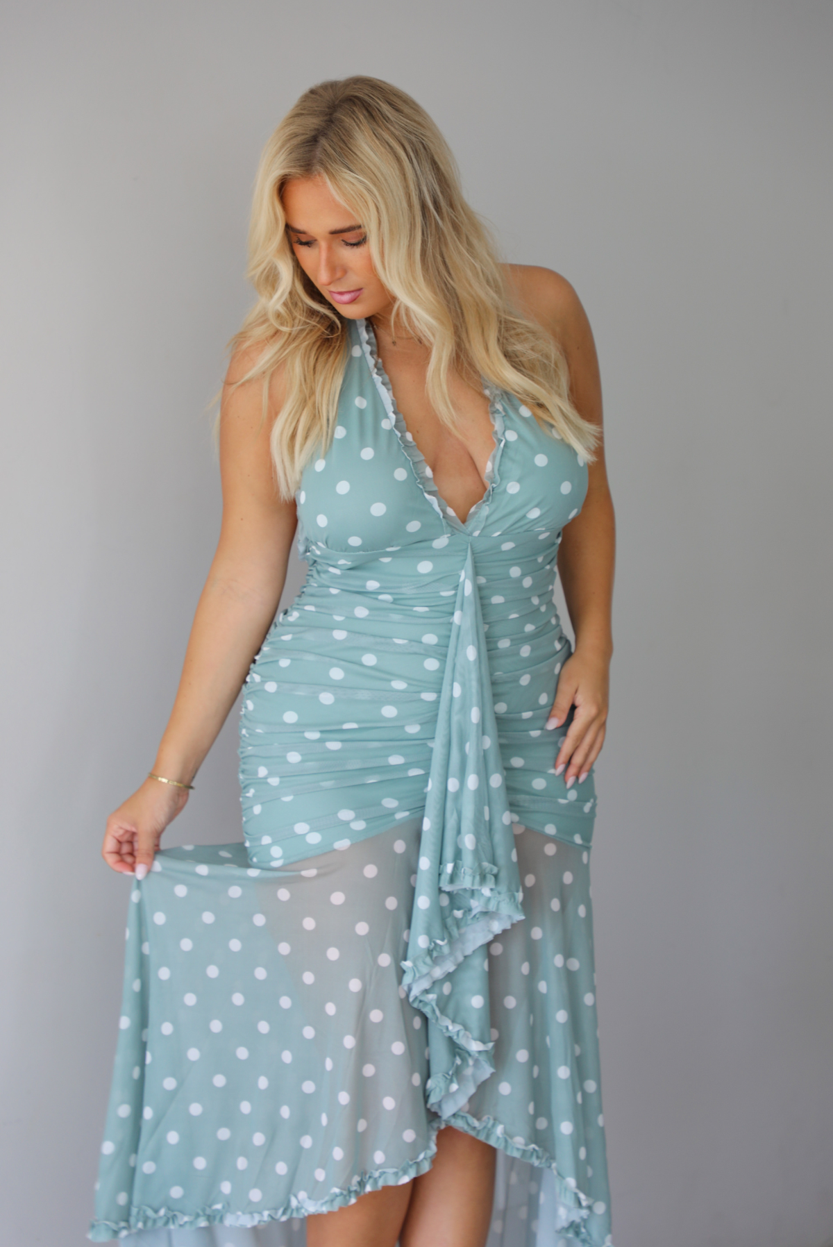 A woman modeling a light sage and white polka dot halter maxi dress with a high-low ruffled hem, standing indoors against a white wall.