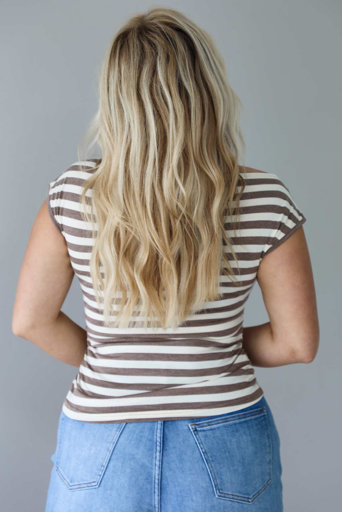Woman wearing a mocha and white striped top with an off the shoulder design with medium wash denim shorts indoors against a white wall. The photo shows the back of the top. 