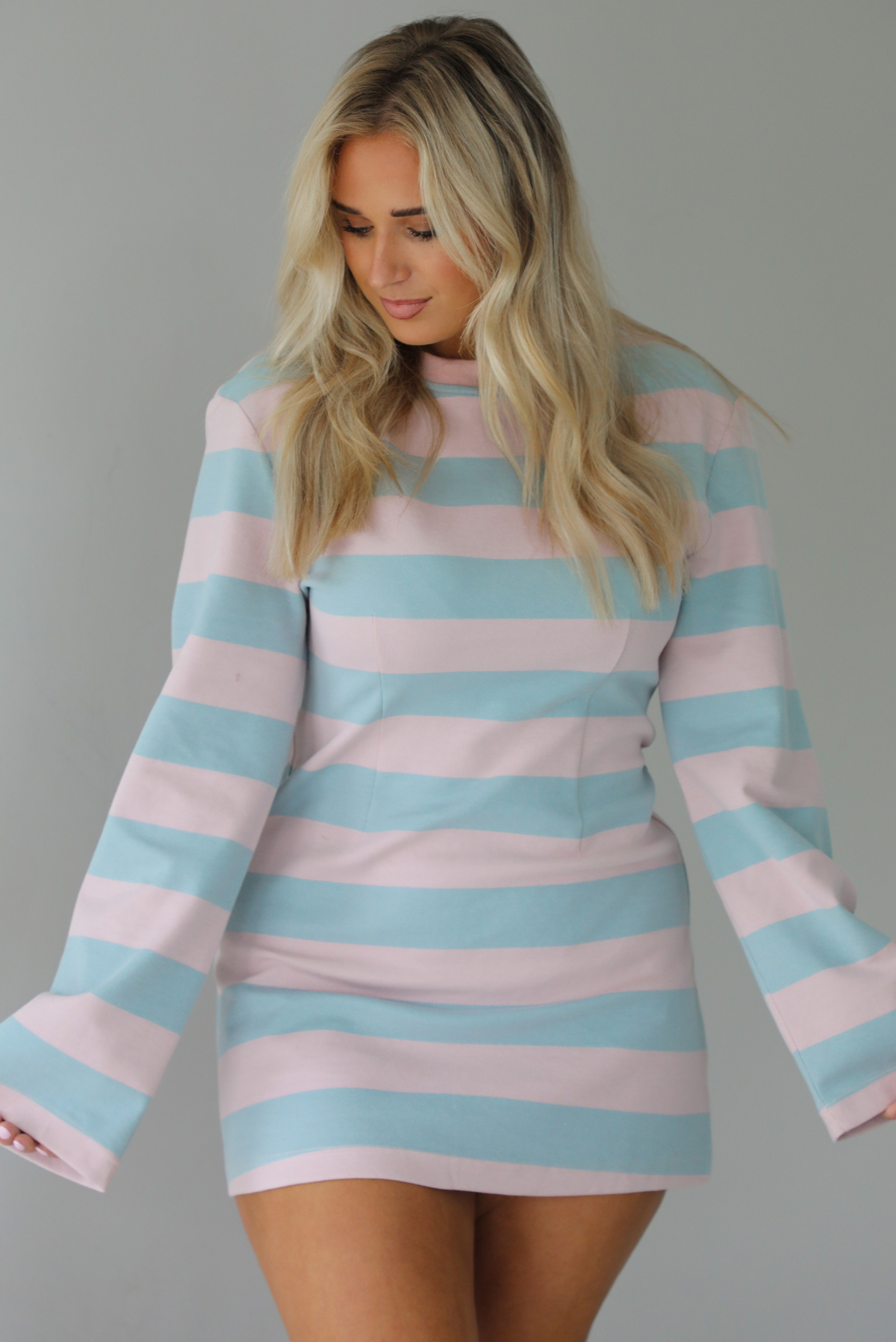 Woman wearing a pink and blue striped dress that has long sleeves and a mini length. The photo is taken indoors against a white wall.