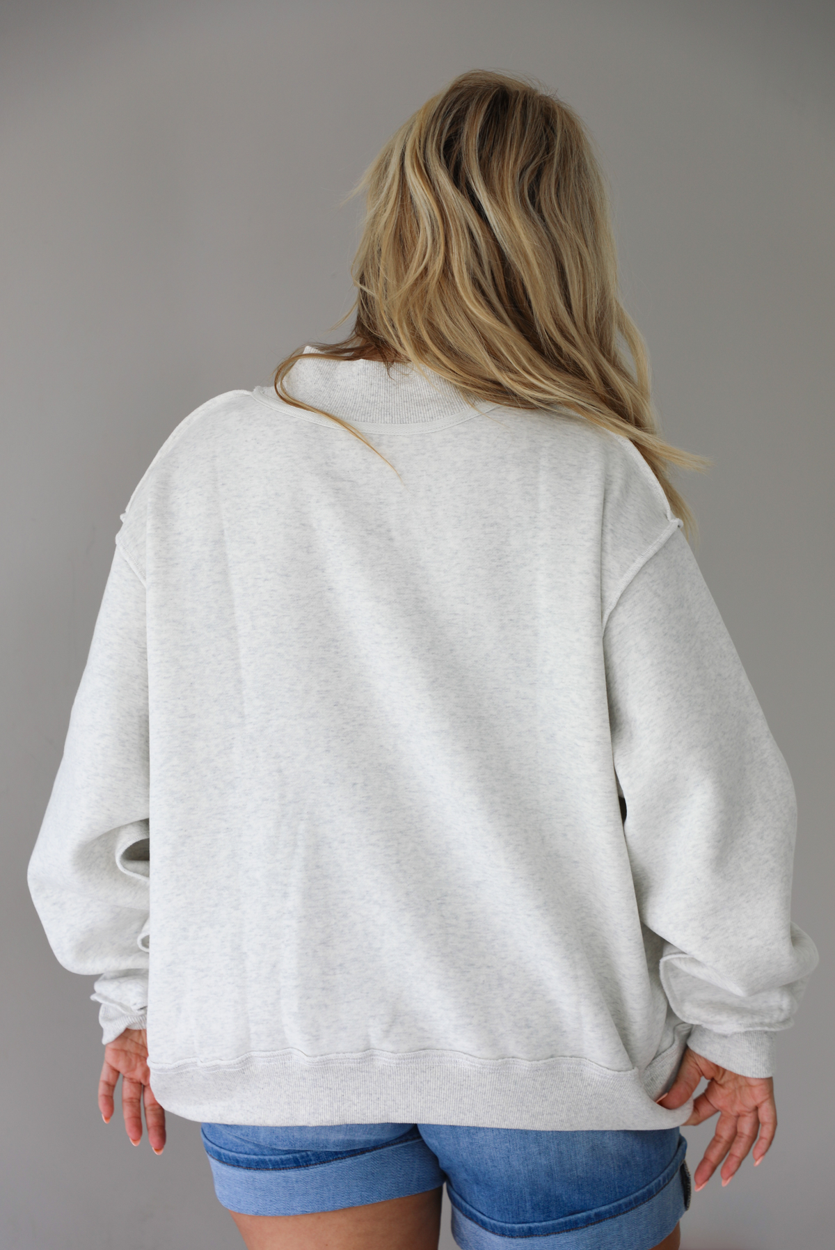 Woman wearing a heather grey oversized sweatshirt, reversed inside out, with the word 'PILATES' written in collegiate font on the front with medium wash denim shorts indoors against a white wall. The photo shows the back of the sweater. 