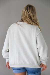 Woman wearing a heather grey oversized sweatshirt, reversed inside out, with the word 'PILATES' written in collegiate font on the front with medium wash denim shorts indoors against a white wall. The photo shows the back of the sweater. 