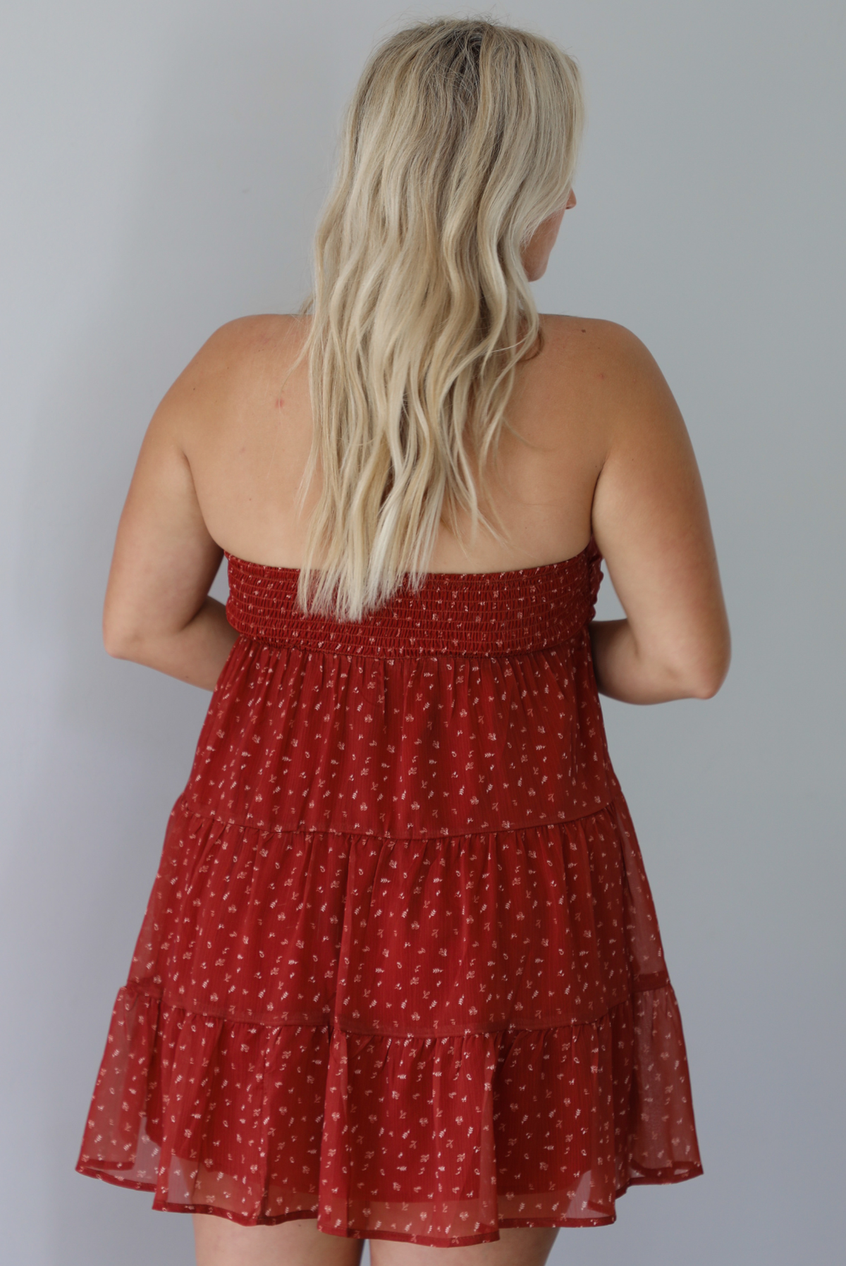 A woman shown from behind wearing a rust mini length dress with a cream floral and leaf tiny print design throughout, has a strapless design with a tie detail in the center of the bust, with a baby-doll dress cut that's fitted at the bust with a flowy skirt, with a smocked detail in the back, standing indoors against a white wall.