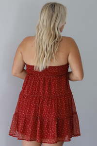 A woman shown from behind wearing a rust mini length dress with a cream floral and leaf tiny print design throughout, has a strapless design with a tie detail in the center of the bust, with a baby-doll dress cut that's fitted at the bust with a flowy skirt, with a smocked detail in the back, standing indoors against a white wall.