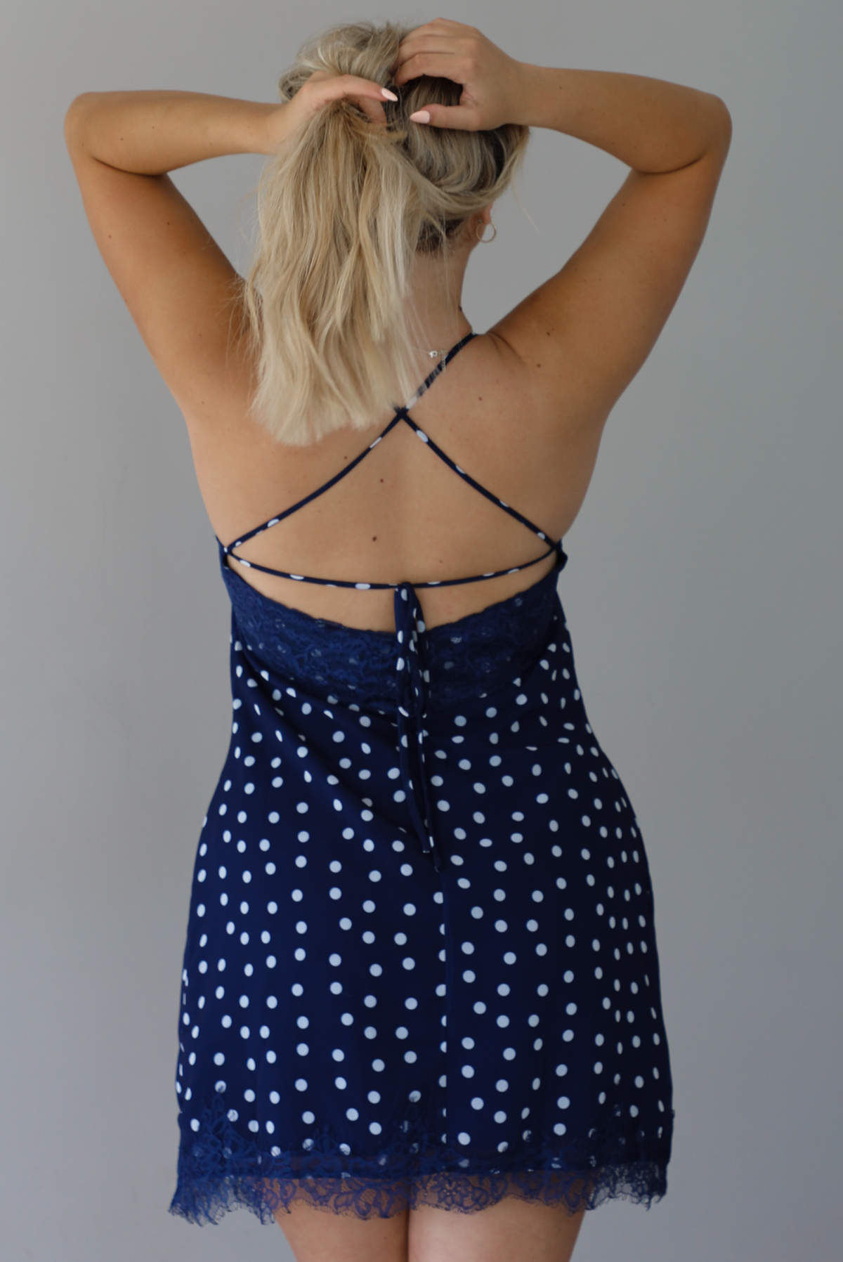Woman wearing a navy dress with a white polka dot print pattern throughout with blue lace detailing at the neckline and hem indoors against a white wall. The photo shows the back of the dress, with its tie detail and zipper closure. 