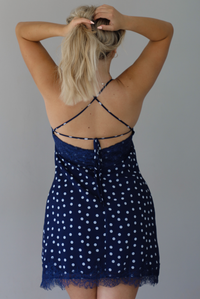 Woman wearing a navy dress with a white polka dot print pattern throughout with blue lace detailing at the neckline and hem indoors against a white wall. The photo shows the back of the dress, with its tie detail and zipper closure. 