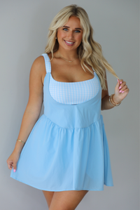 A woman modeling a light blue sleeveless mini dress with a white gingham bodice and a flared skirt, paired with brown slide sandals, standing indoors against a white wall.
