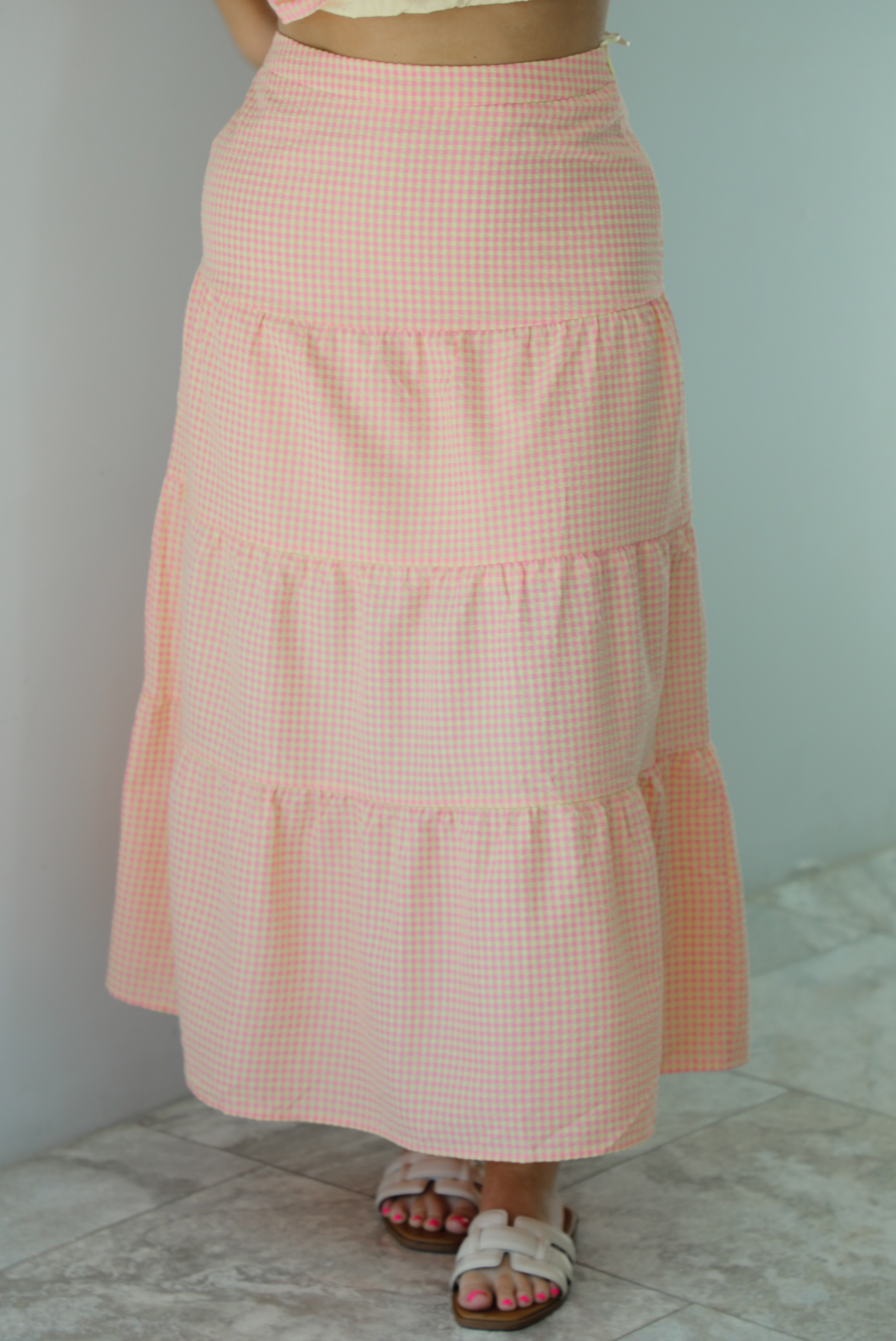 Woman wearing a pink and pale yellow gingham print two piece set featuring a smocked top and high waisted, maxi length skirt. The photo is taken indoors against a white wall and shows a closeup of the skirt, which is high waisted and has a maxi length.
