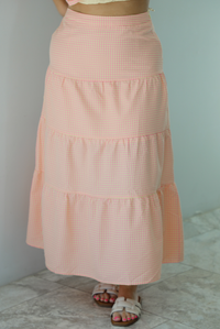 Woman wearing a pink and pale yellow gingham print two piece set featuring a smocked top and high waisted, maxi length skirt. The photo is taken indoors against a white wall and shows a closeup of the skirt, which is high waisted and has a maxi length.