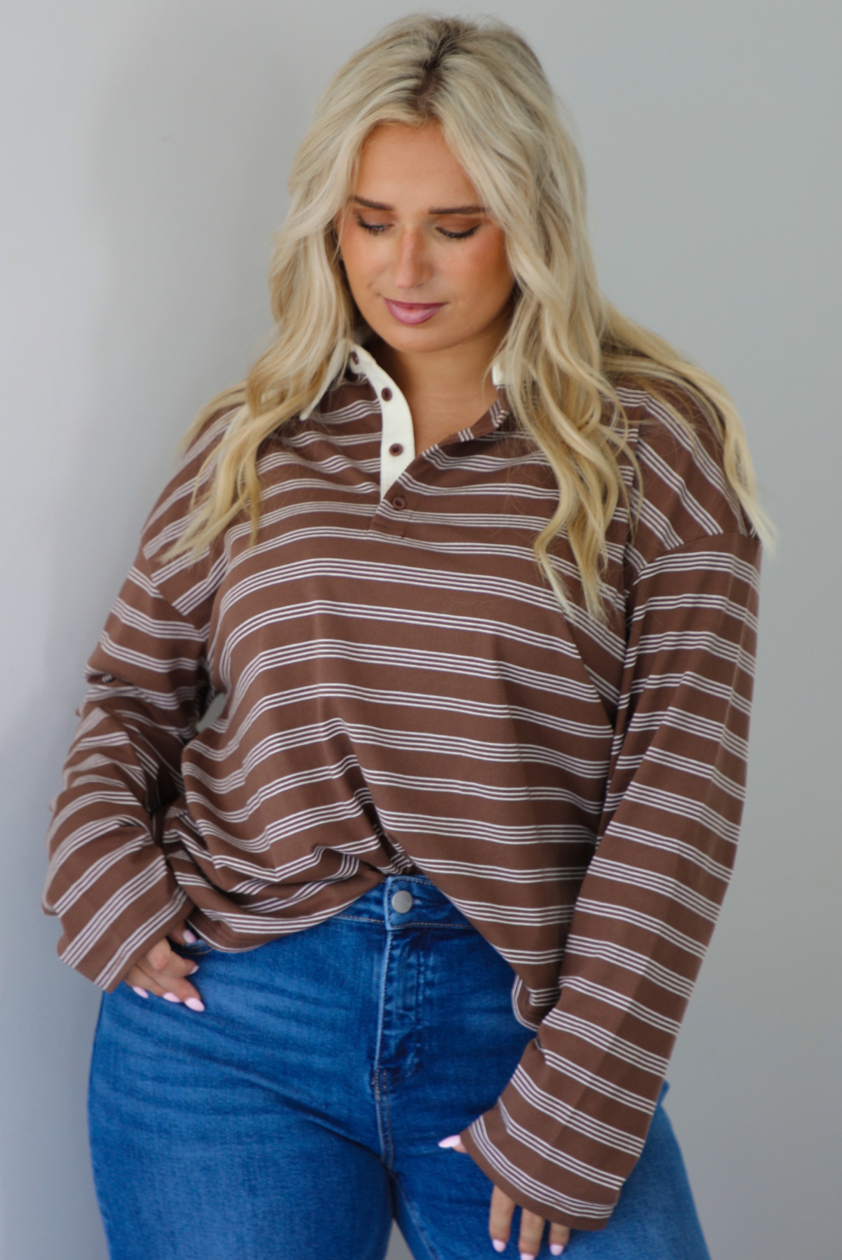 A woman wearing a brown and cream striped collared quarter-button long sleeved top, front tucked into dark wash denim wide leg jeans, standing indoors against a white wall.