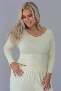 Woman wearing a light yellow three piece set featuring a long sleeved top, long, flared hem pants, and a long, matching cardigan. The photo is taken indoors against a white wall and shows a closeup of the top. 